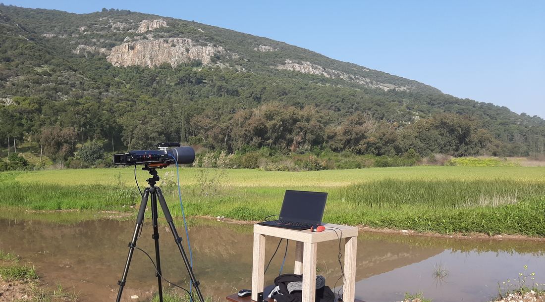 A small telescope with a camera on a tripod in a field, pointed at a wooded hillside, with a laptop on a table beside it.