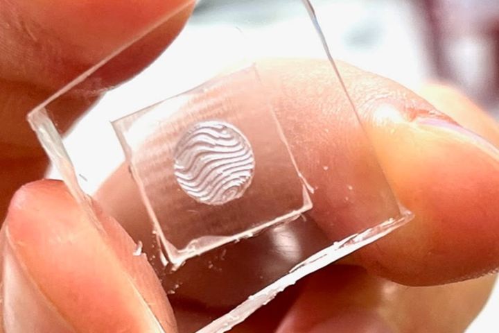 Fingers holding a small clear plate with a round patch of wavy relief at its center.