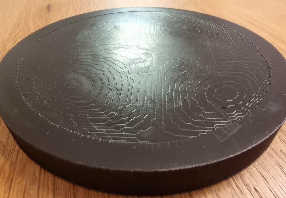 A dark disc lying on a wooden table. Its top face carries a relief of stepped contour lines, like a terraced map.